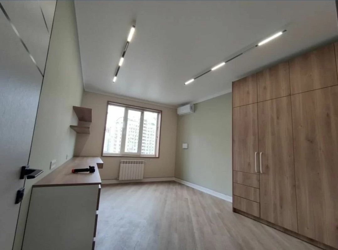 Jasne wnętrze pokoju po remoncie: drewniana podłoga, szafa w zabudowie, biurko z półkami, grzejnik pod oknem i nowoczesne oświetlenie sufitowe.