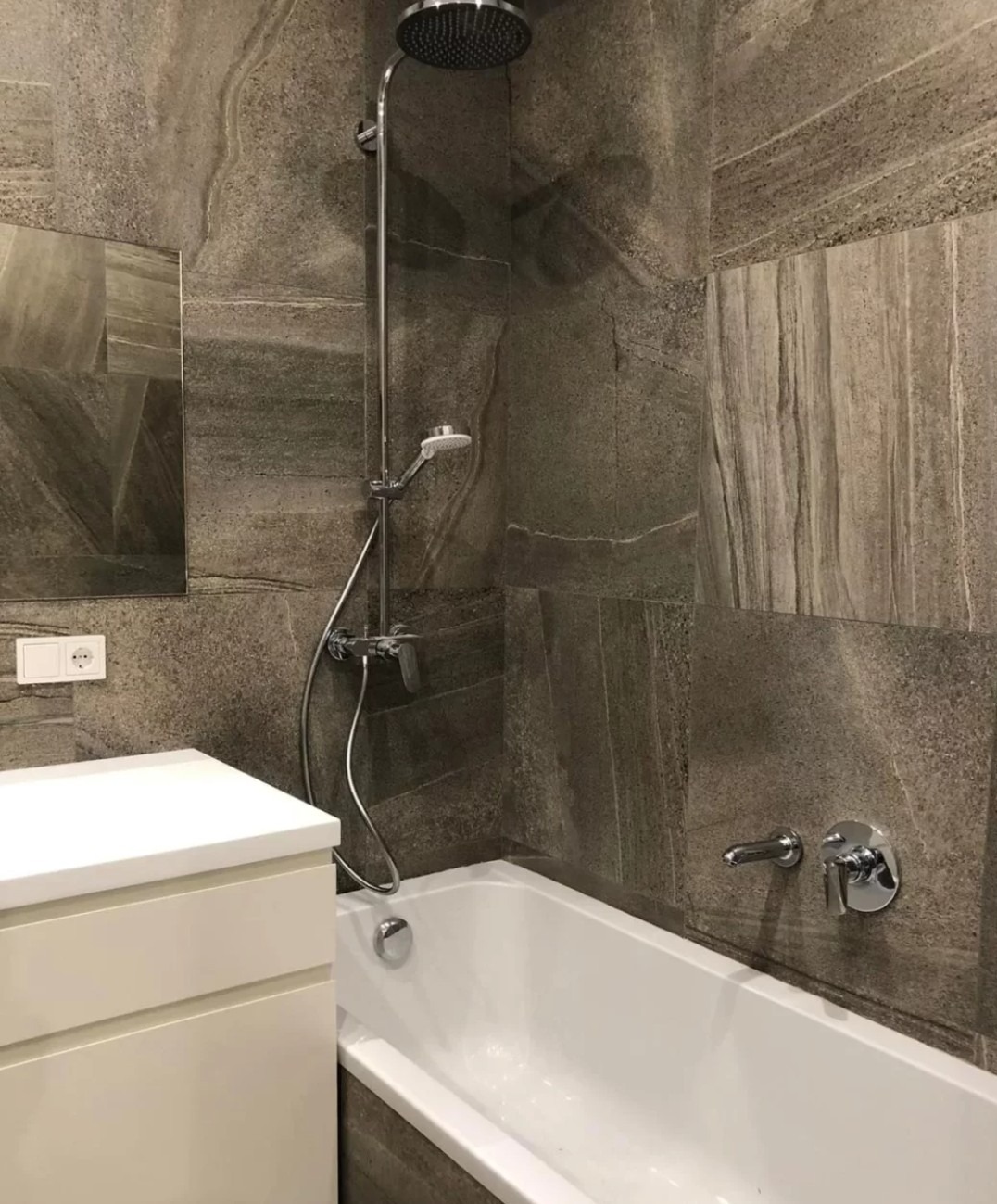 Nowoczesna łazienka z wanną, deszczownicą i umywalką w zabudowie. Ciemne płytki imitujące kamień na ścianach. Minimalistyczny styl.