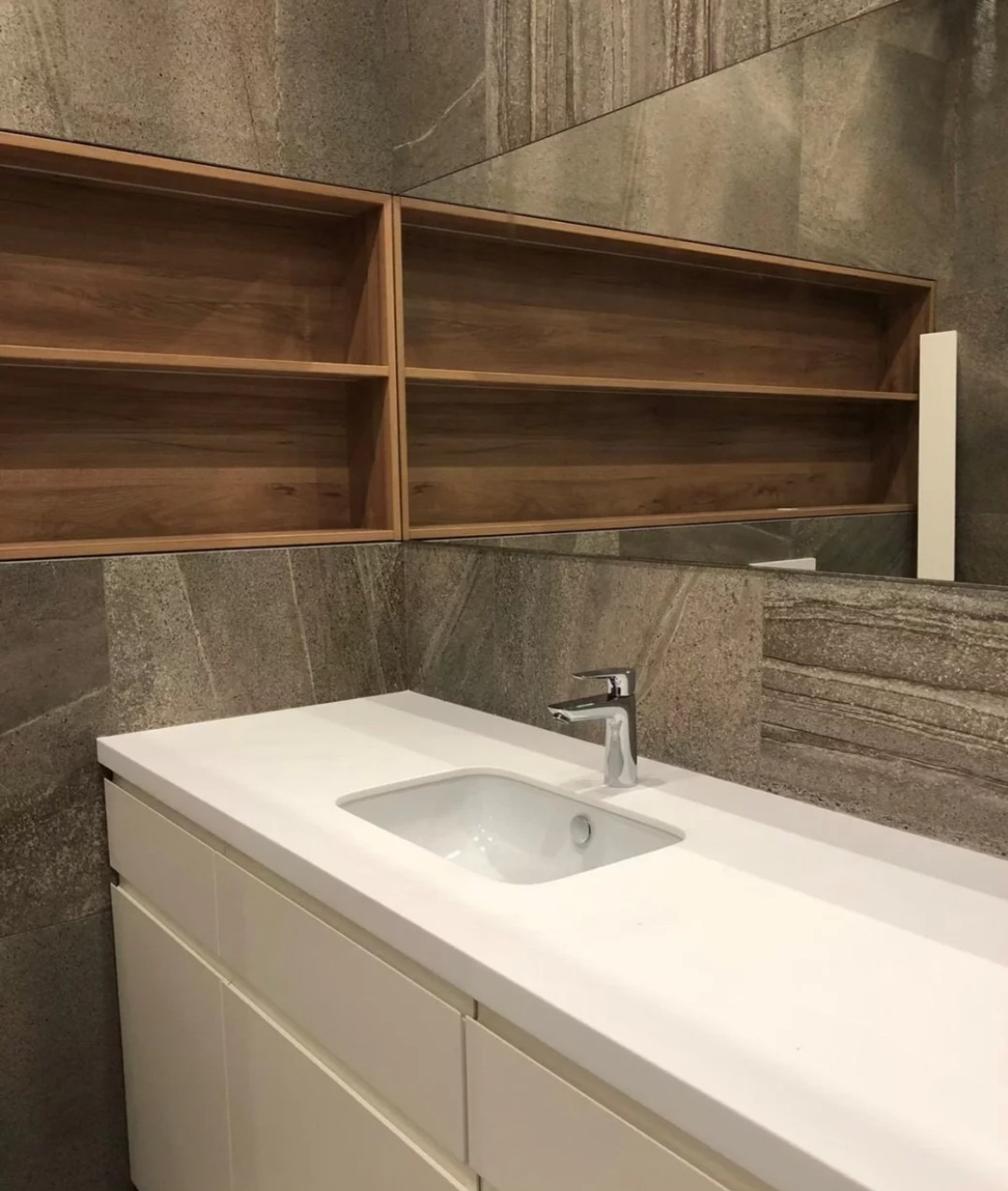 Nowoczesna łazienka z białą umywalką nablatową, drewnianymi półkami i płytkami imitującymi kamień. Bateria chromowana. Minimalistyczny design.