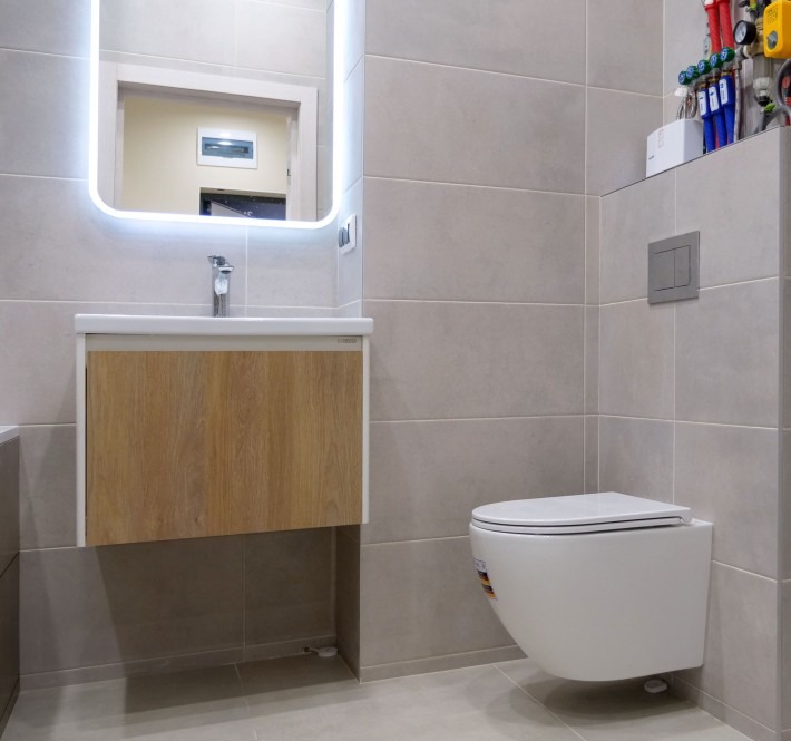 Nowoczesna łazienka z podwieszaną toaletą, umywalką z szafką o drewnianym froncie i podświetlanym lustrem. Szare płytki na ścianach i podłodze.