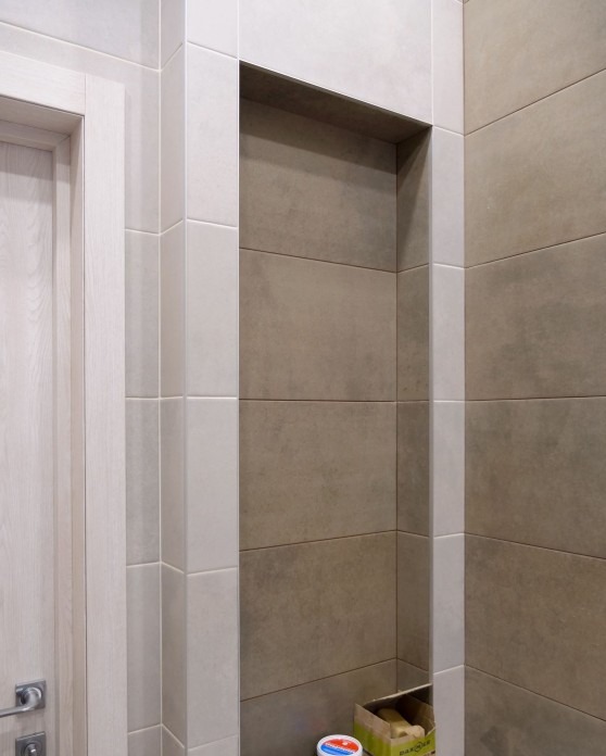 Wykończona wnęka w łazience z szarymi płytkami. Widoczna część drzwi i narzędzia w kartonie na dole. Minimalistyczny design.