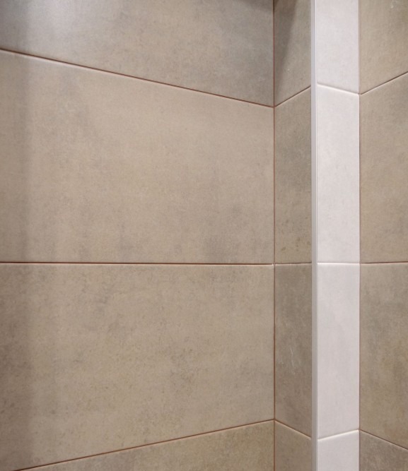 Beżowe, matowe płytki ceramiczne ułożone poziomo, z wąską, pionową kolumną białych płytek w rogu. Widoczne fugi w odcieniach brązu.