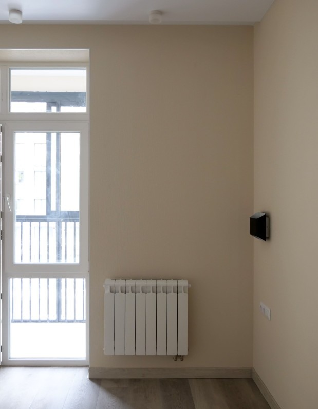 Jasne wnętrze po remoncie: beżowe ściany, biały grzejnik pod oknem balkonowym, drewnopodobna podłoga. Minimalistyczny design, czysty kadr.