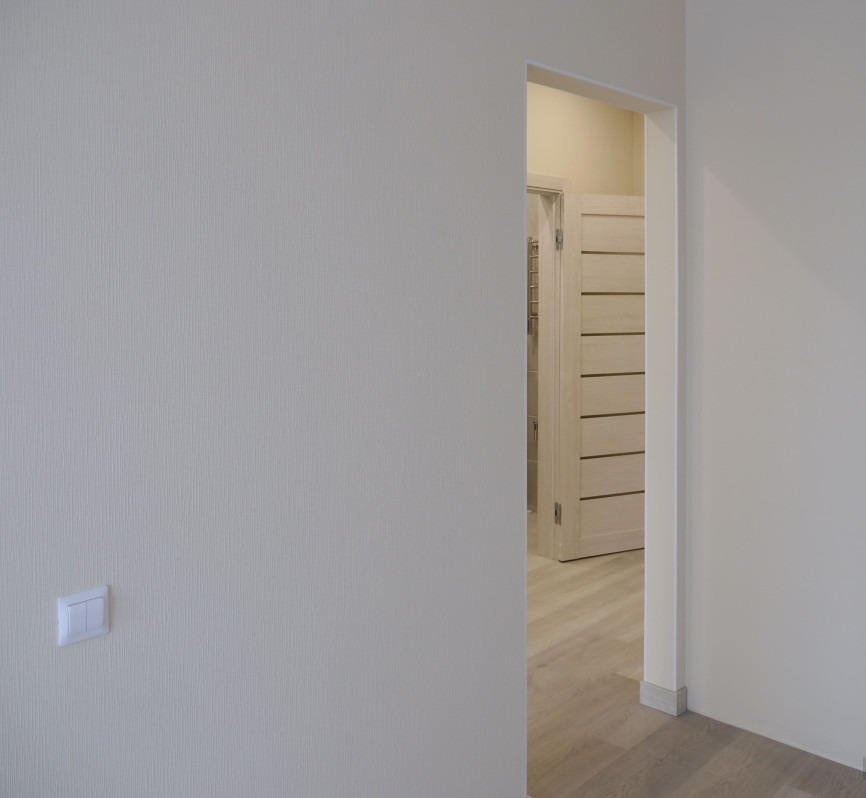 Jasny pokój po remoncie: kremowe ściany z delikatną fakturą, biały włącznik światła, widok na drzwi z poziomymi panelami i podłogę w odcieniach beżu.