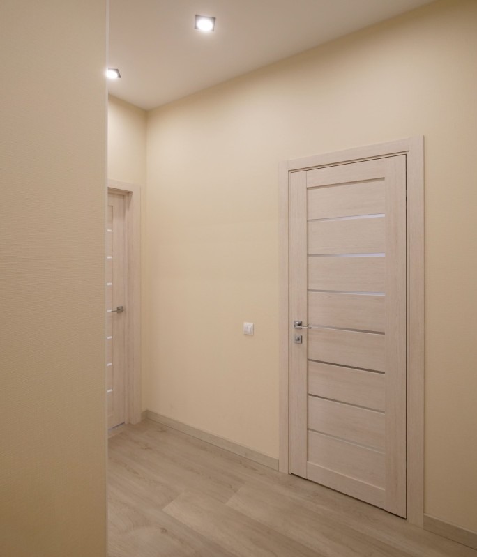 Jasny korytarz po remoncie w Katowicach: kremowe ściany, drewnopodobna podłoga i dwie białe, nowoczesne drzwi z poziomymi przeszkleniami.