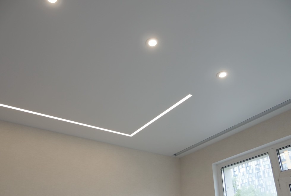 Nowoczesny sufit podwieszany z oświetleniem LED w kształcie litery L i trzema punktowymi lampami, jasne ściany, okno w prawym dolnym rogu.