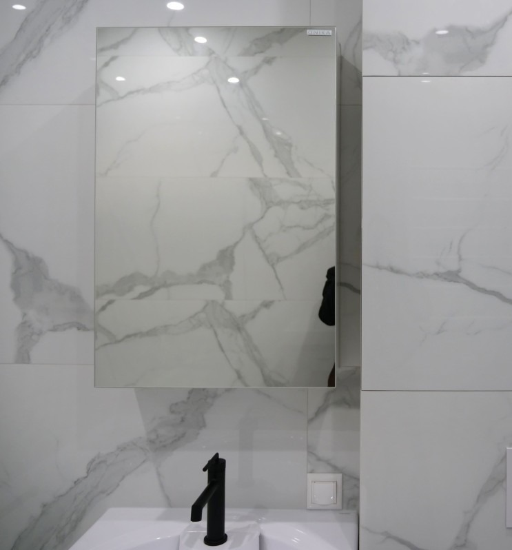 Biała łazienka z płytkami imitującymi marmur, czarna bateria umywalkowa i lustro z szafką, odbijające fragment wnętrza. Minimalistyczny styl.