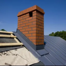 Ceglany komin na dachu krytym blachą w Warszawie, widoczne drewniane elementy konstrukcji i obróbki blacharskie.
