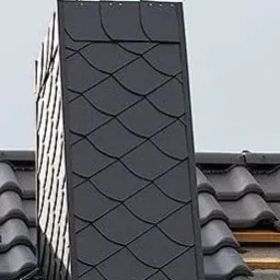 Szary komin obłożony łuską dachową, widoczny na tle dachu z dachówki ceramicznej, częściowo w trakcie krycia. Zastosowano metalowy wspornik kominiarski.