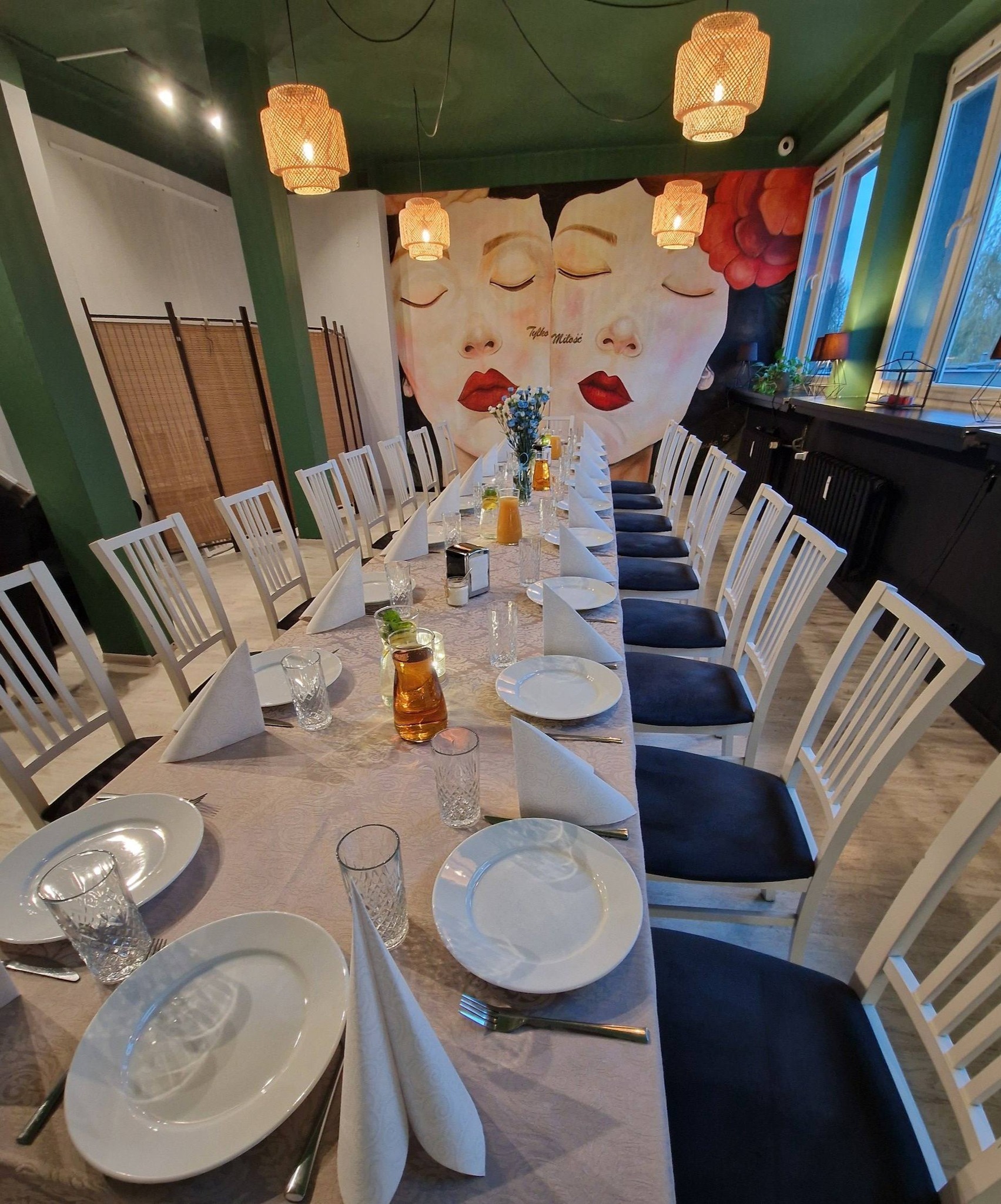 Długi stół z nakryciami, białymi talerzami i szklankami, ustawiony w restauracji z muralem przedstawiającym twarze i rattanowymi lampami.