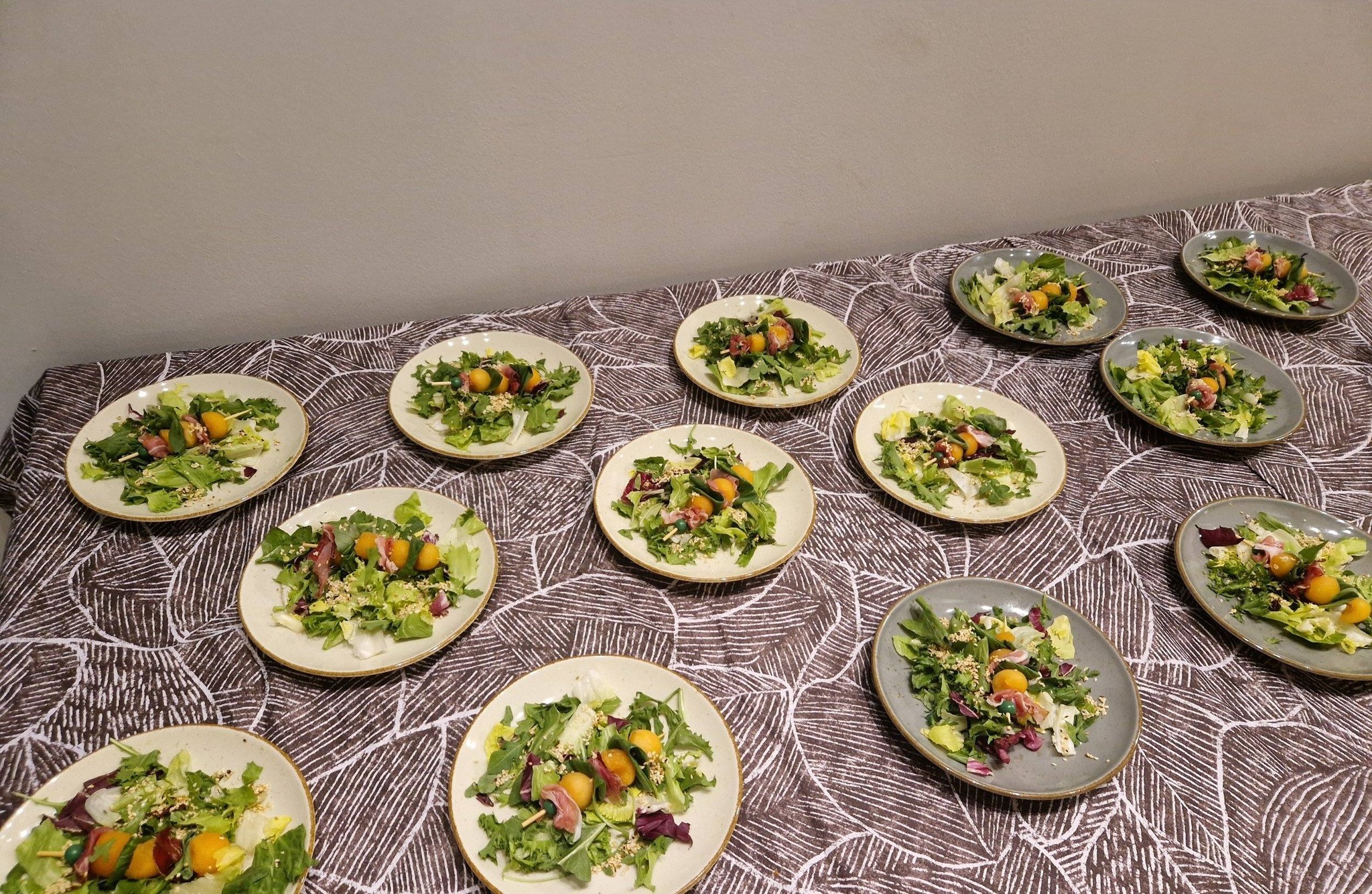 Ustawione na stole talerze z sałatkami, dekorowane melonem i szynką, gotowe do serwowania. Talerze na brązowym obrusie w geometryczny wzór.