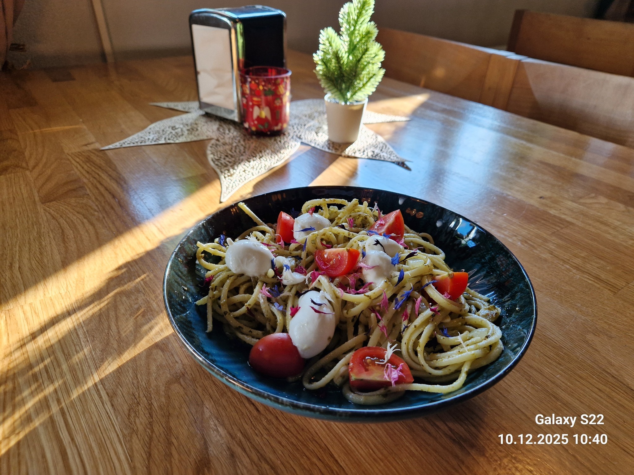 Apetyczne spaghetti z pesto, pomidorkami i mini mozzarellą na ciemnym talerzu. W tle dekoracje stołu: serwetnik, świecznik i mała choinka. Ciepłe światło podkreśla detale.