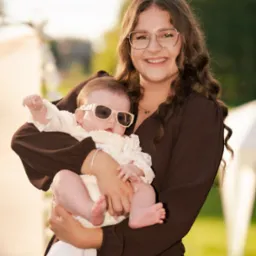 Uśmiechnięta kobieta w brązowej sukience trzyma na rękach niemowlę w białym ubranku i okularach przeciwsłonecznych na tle zieleni.