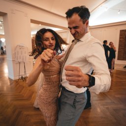 Cherrytime - Para w tańcu na parkiecie podczas wesela. Kobieta w błyszczącej sukni, mężczyzna w koszuli i spodniach. W tle stoliki z gośćmi i dekoracje.