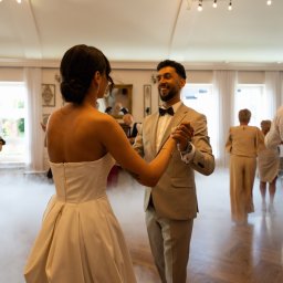 Cherrytime - Para młoda tańczy pierwszy taniec w sali balowej z dekoracją świetlną i efektem dymu. Pan młody w beżowym garniturze, panna młoda w sukni bez ramiączek.