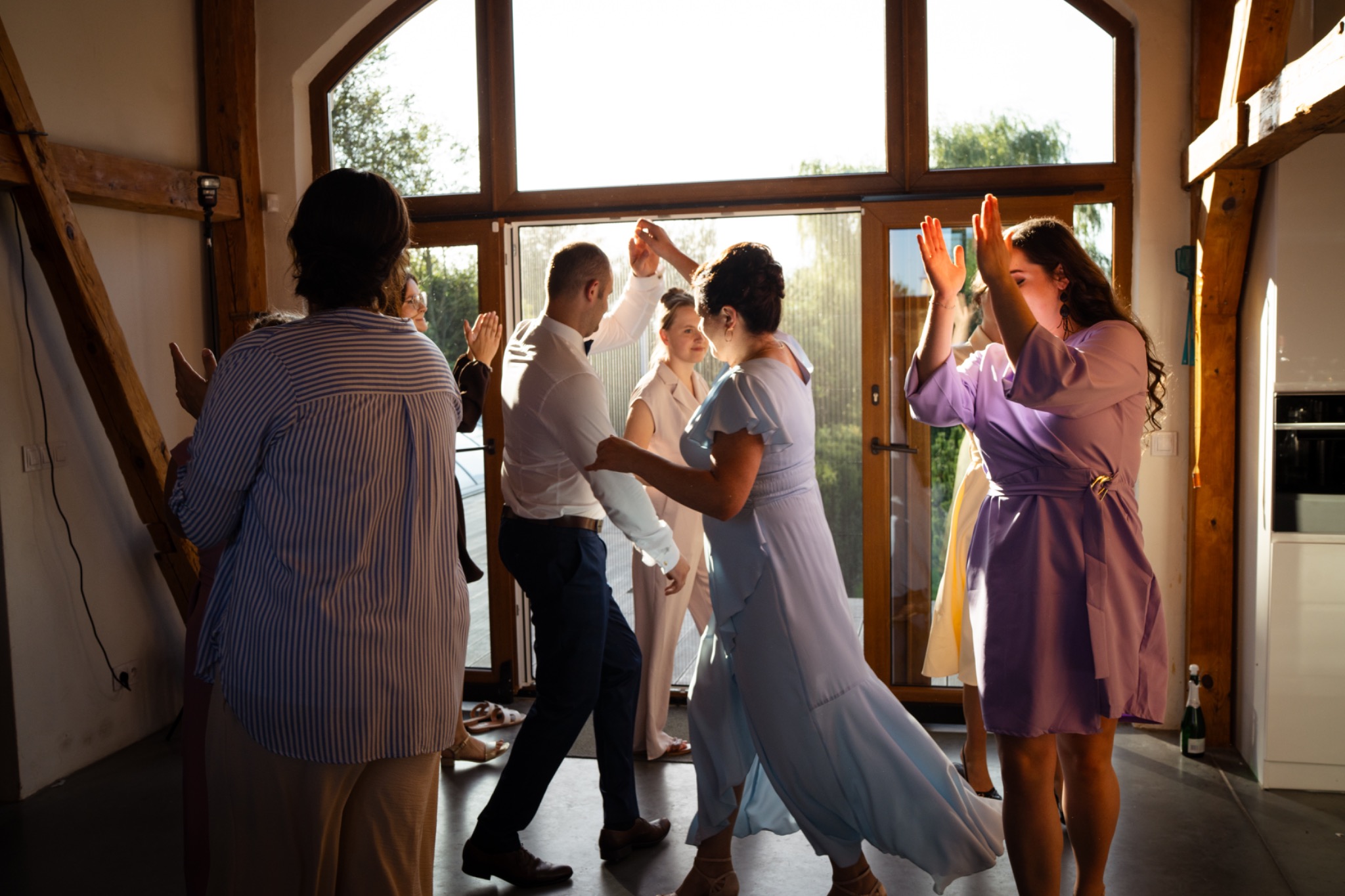 Goście weselni tańczą w jasnym pomieszczeniu z dużym oknem, w tle widoczna zieleń. Oświetlenie słoneczne, radosna atmosfera.