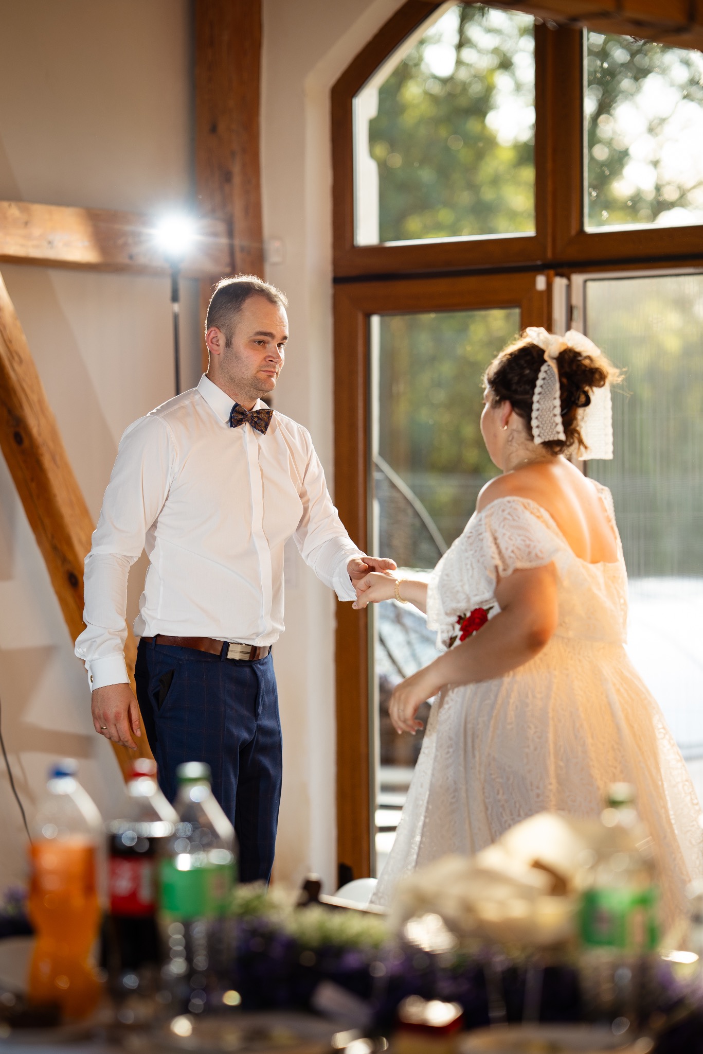 Para młoda trzymająca się za ręce podczas wesela w jasnym wnętrzu z drewnianymi elementami, widoczne butelki na stole.