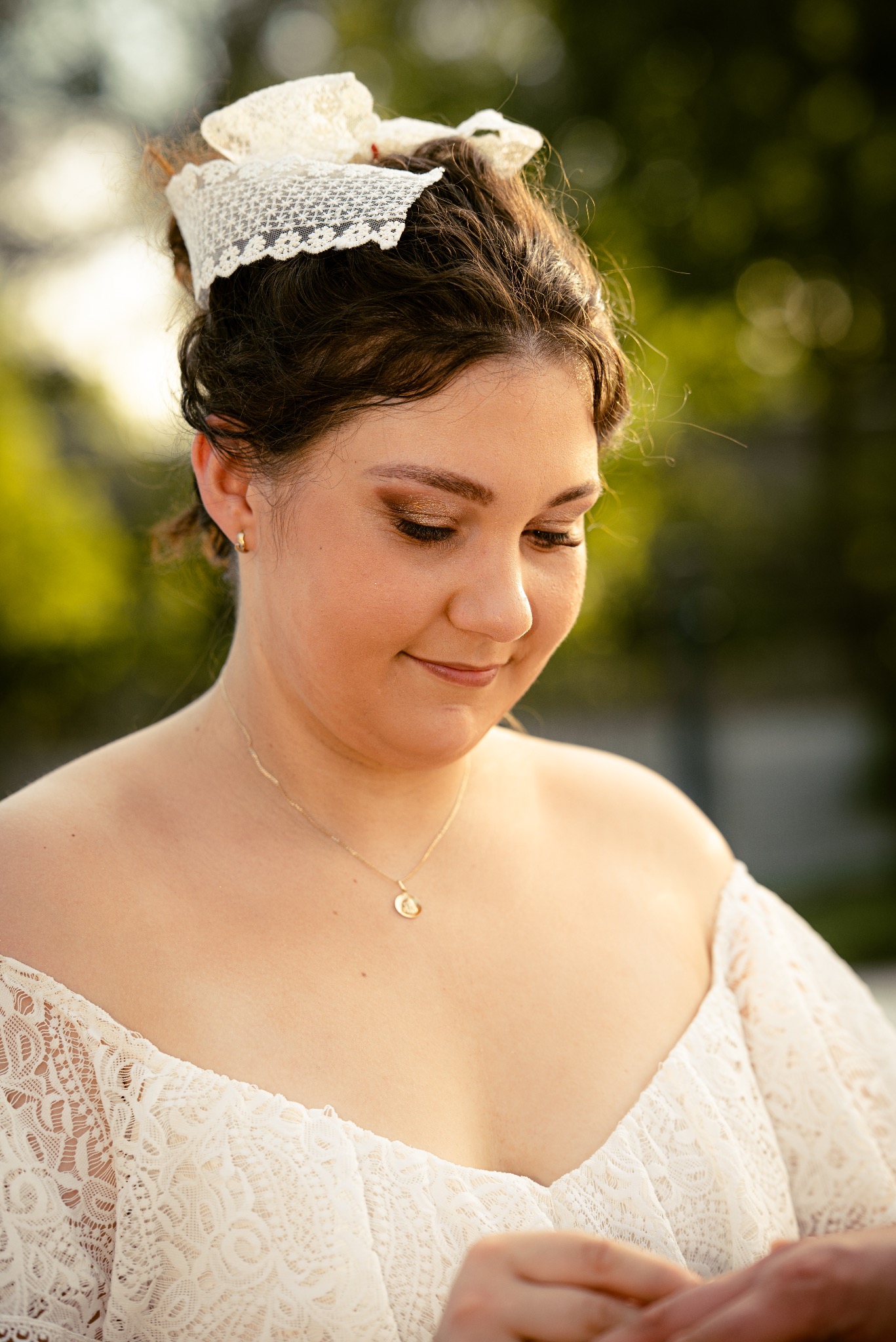 Panna młoda w koronkowej sukni spogląda w dół, na palcu błyszczy obrączka. Delikatny makijaż i złoty łańcuszek dopełniają stylizację. Włosy upięte w kok z koronkową ozdobą.