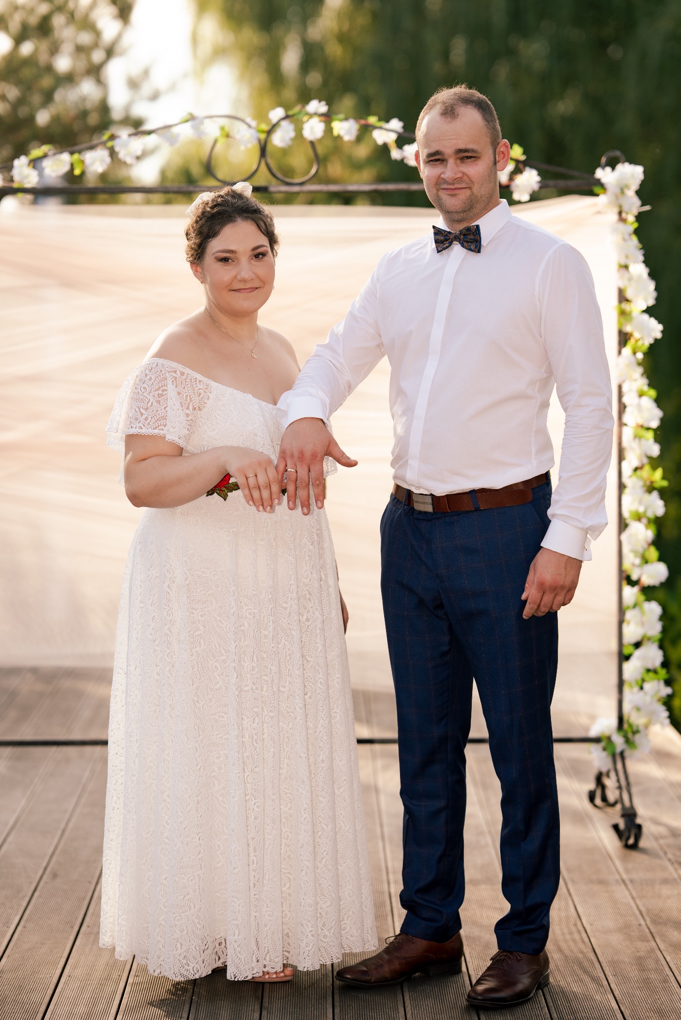 Para młoda prezentuje obrączki na tle kwiatowej dekoracji. Panna młoda w koronkowej sukni, pan młody w muszce i wzorzystych spodniach. Romantyczna atmosfera.