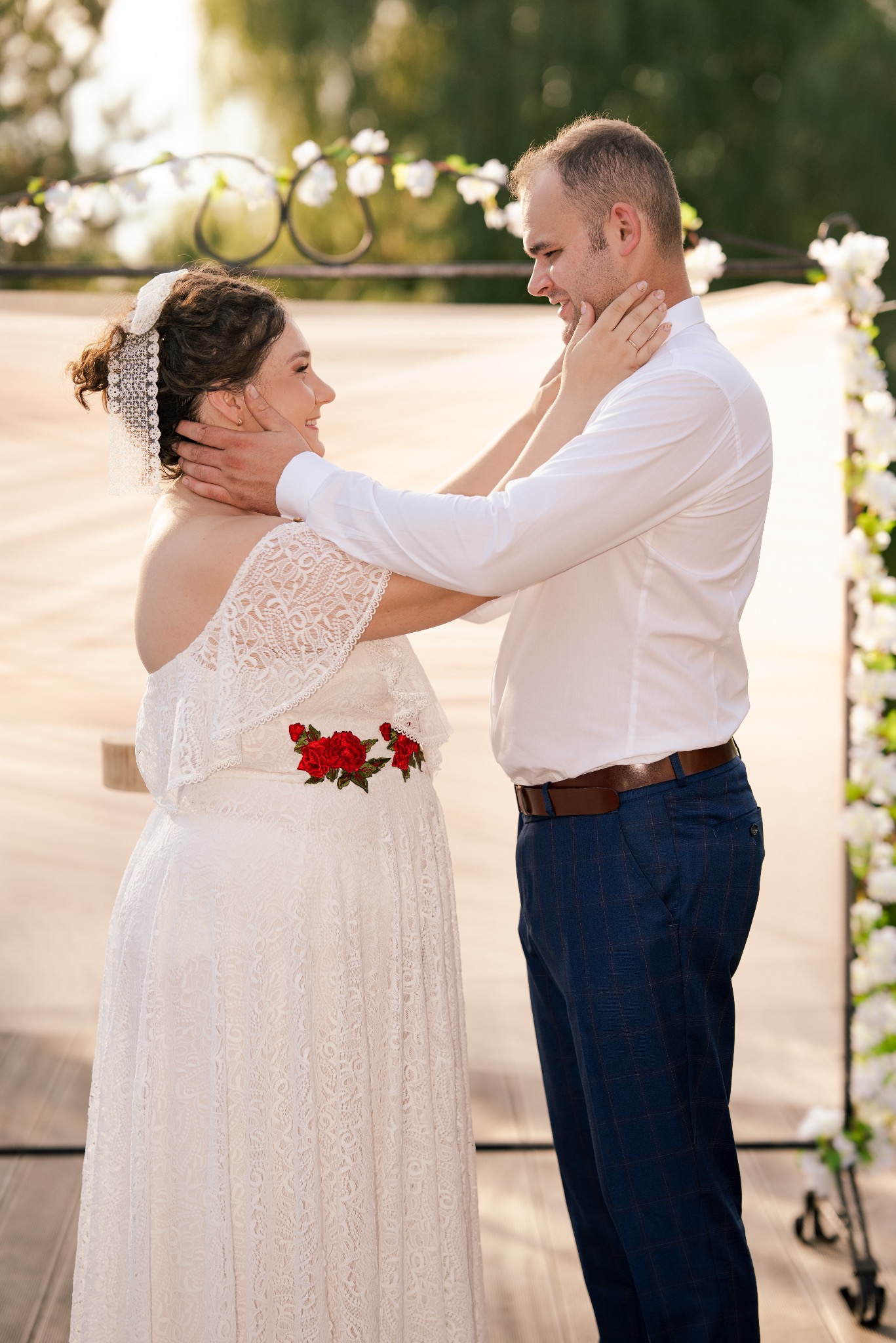 Para młoda w objęciach przed udekorowanym kwiatami łukiem. Panna młoda w koronkowej sukni z czerwonymi różami, pan młody w niebieskich spodniach w kratę.