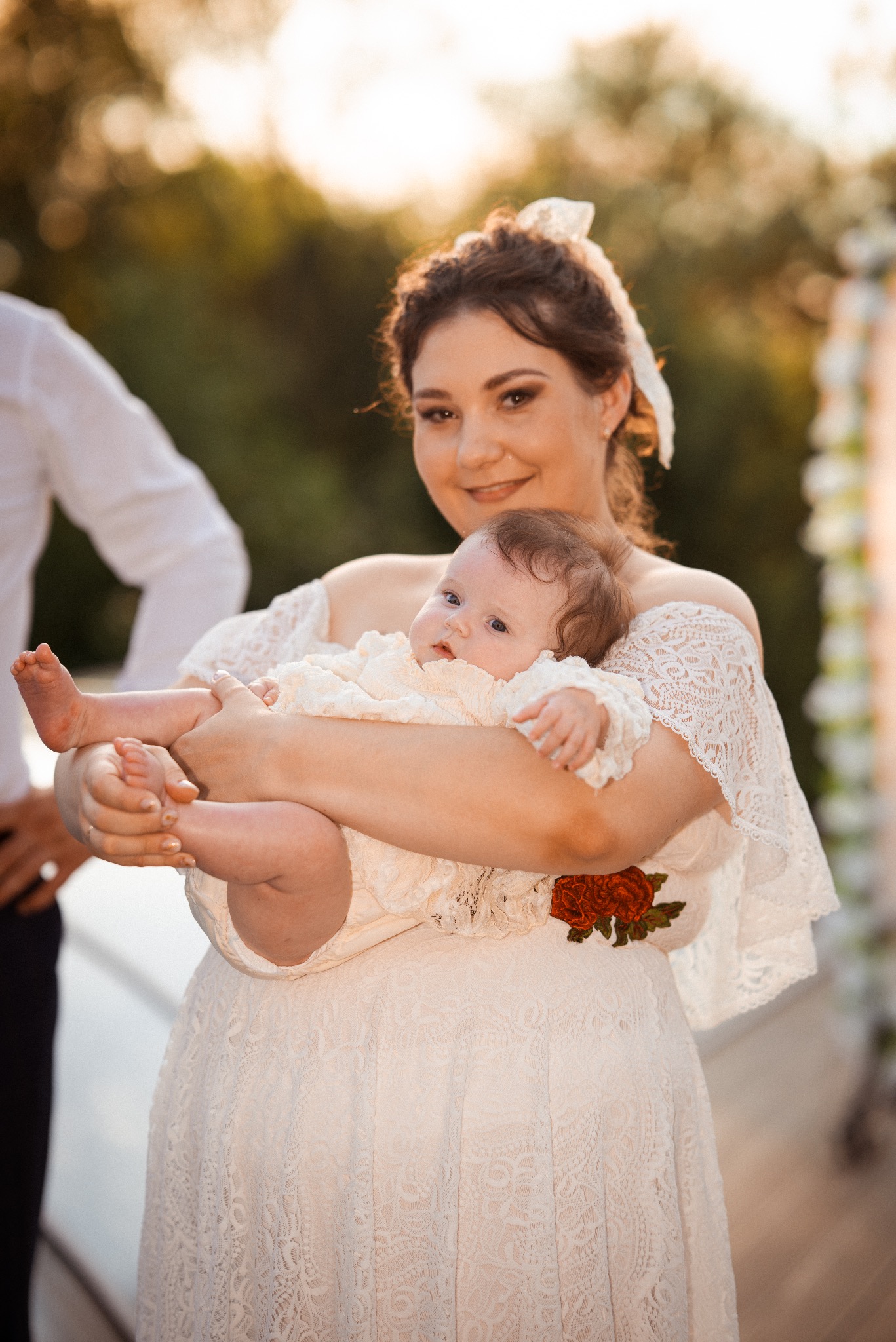 Uśmiechnięta kobieta w koronkowej sukni trzyma niemowlę na tle dekoracji ślubnej. Ciepłe, naturalne światło.