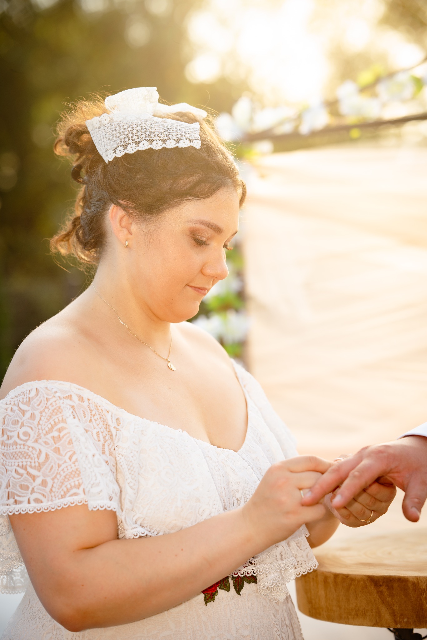 Panna młoda w koronkowej sukni zakłada obrączkę na palec pana młodego podczas ceremonii ślubnej w plenerze, oświetlonej ciepłym, naturalnym światłem.