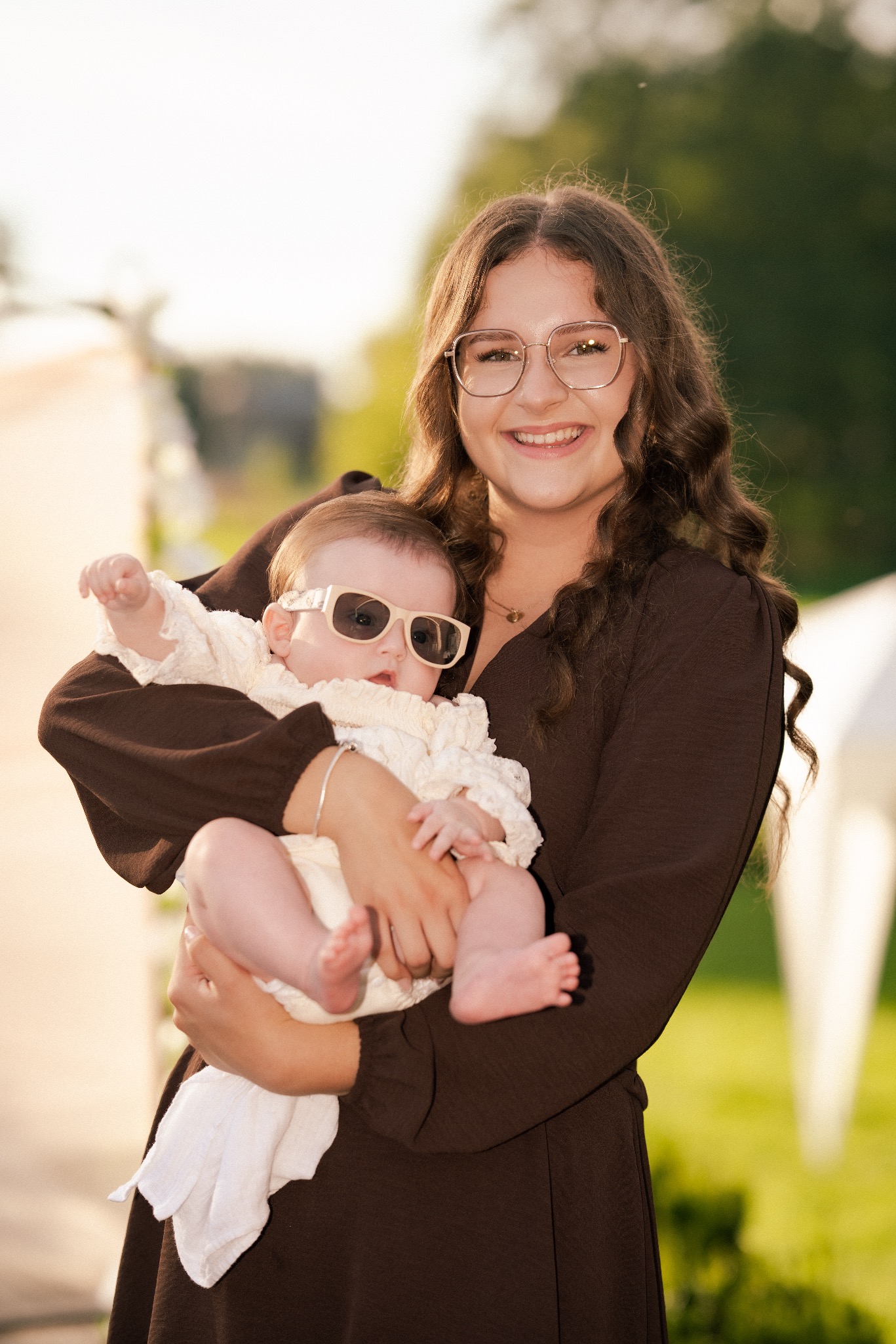Uśmiechnięta kobieta w brązowej sukience trzyma na rękach niemowlę w białym ubranku i okularach przeciwsłonecznych na tle zieleni.