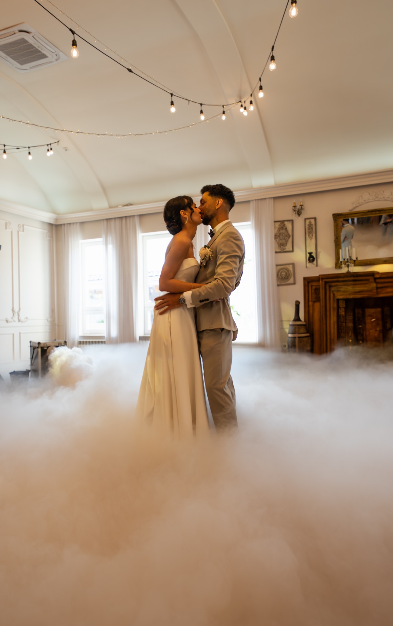 Pocałunek pary młodej w tanecznym obłoku mgły, dekoracyjne oświetlenie, jasne wnętrze z kominkiem w tle. Ujęcie z uroczystości weselnej.