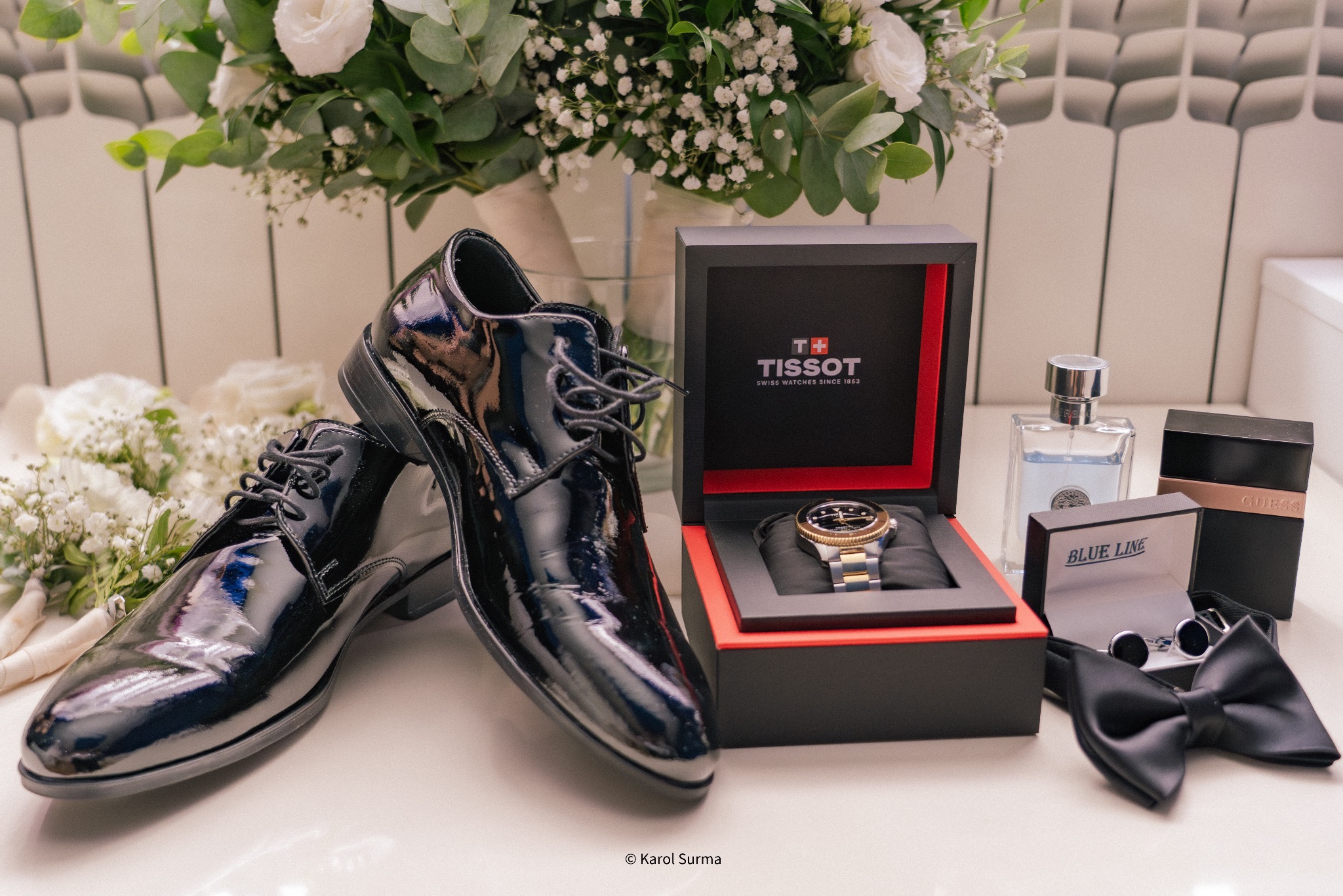 Eleganckie buty ślubne lakierki obok bukietu, zegarka Tissot w pudełku, perfum i spinek do mankietów, tworzące stylową kompozycję dla pana młodego.