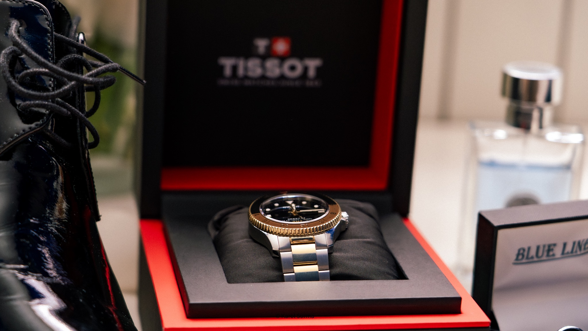Zegarek Tissot w pudełku na czarnej poduszce, obok lakierowany but i flakon perfum Blue Line. Precyzyjne detale i eleganckie tło tworzą unikalną kompozycję.