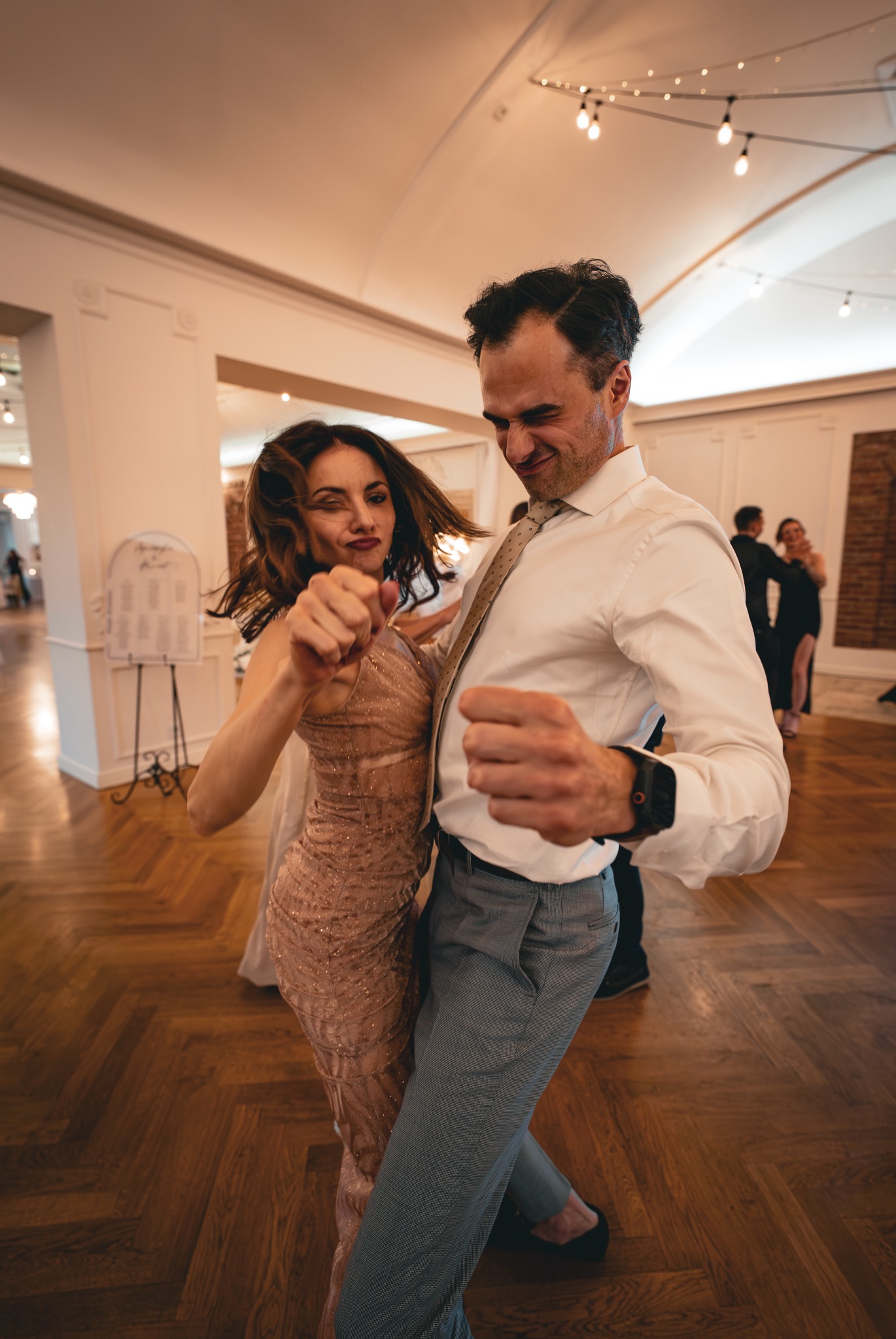 Para w tańcu na parkiecie podczas wesela. Kobieta w błyszczącej sukni, mężczyzna w koszuli i spodniach. W tle stoliki z gośćmi i dekoracje.