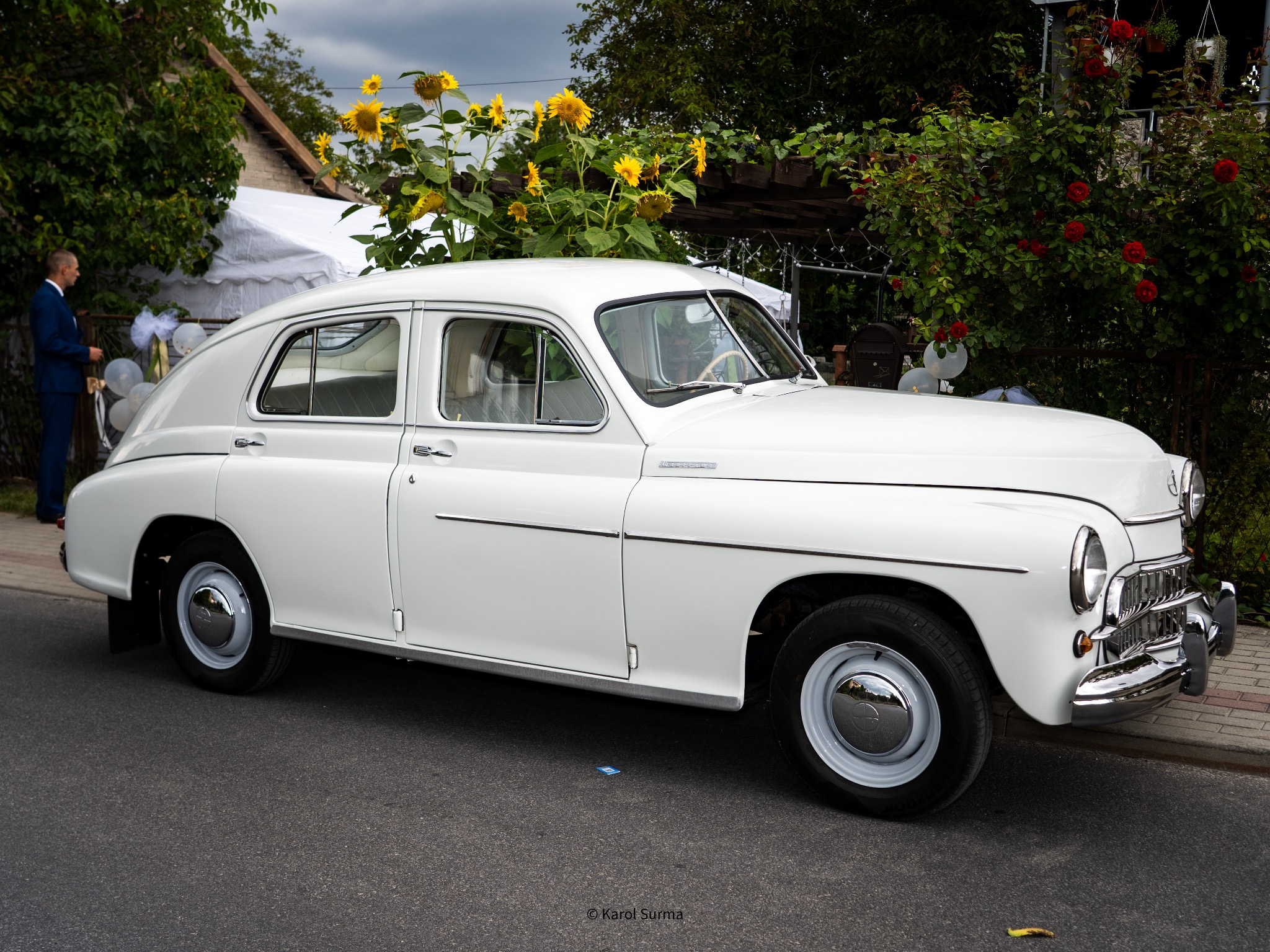 Elegancki, biały samochód GAZ-21 Wołga, udekorowany na ślub, zaparkowany na ulicy z dekoracjami z kwiatów i balonów w tle. Klasyczny pojazd na uroczystość.