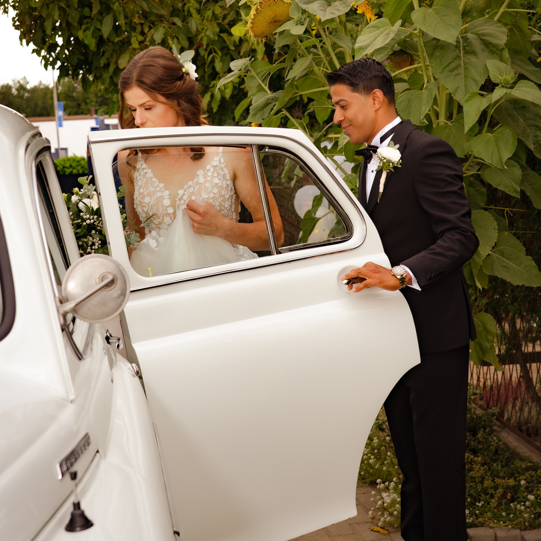 Panna młoda w koronkowej sukni wsiada do białego, zabytkowego samochodu. Pan młody w smokingu otwiera drzwi, w tle słoneczniki.