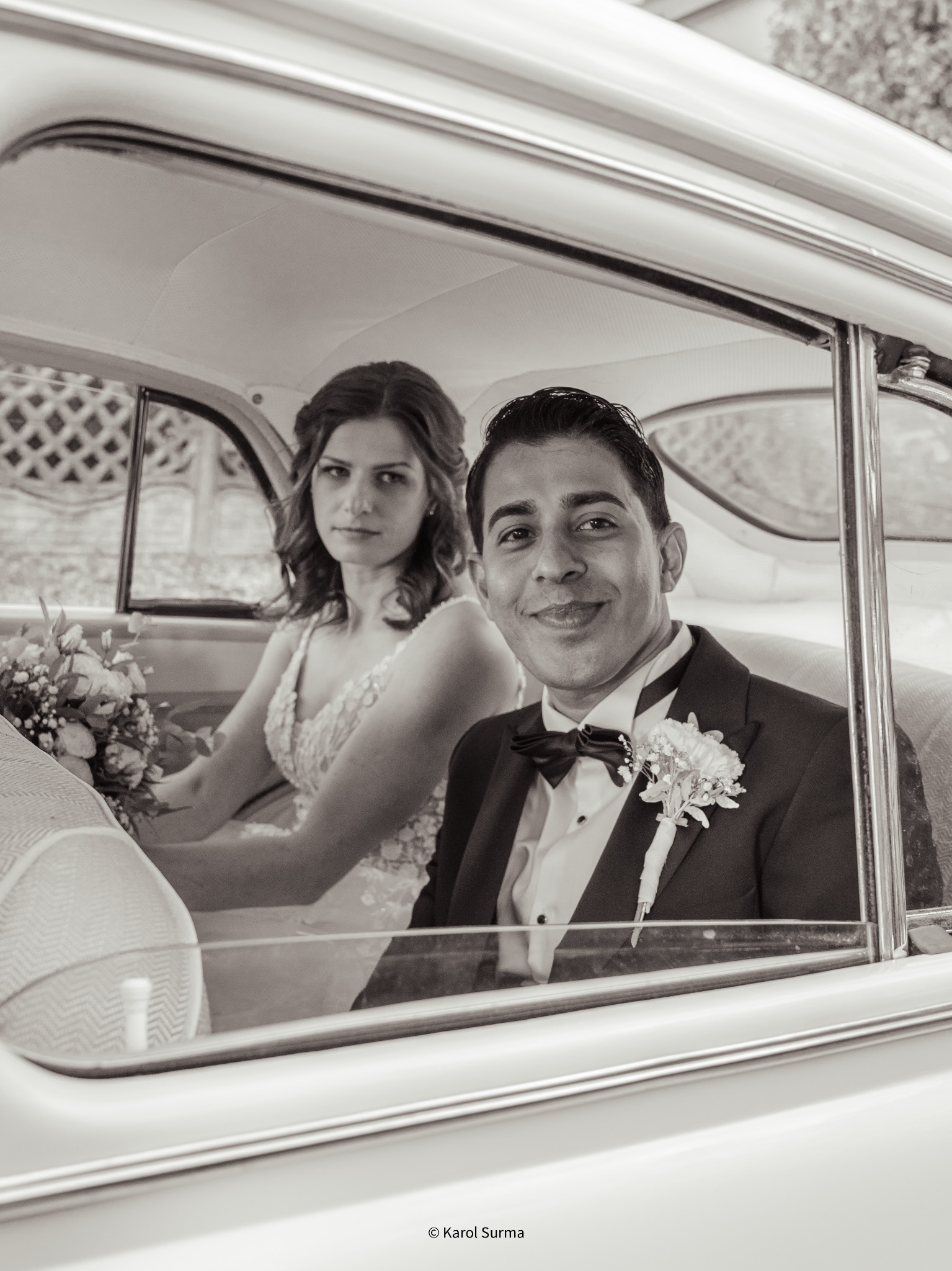 Para młoda w zabytkowym samochodzie: pan młody w smokingu z butonierką uśmiecha się, panna młoda z bukietem spogląda poważnie. Czarno-biała fotografia ślubna.