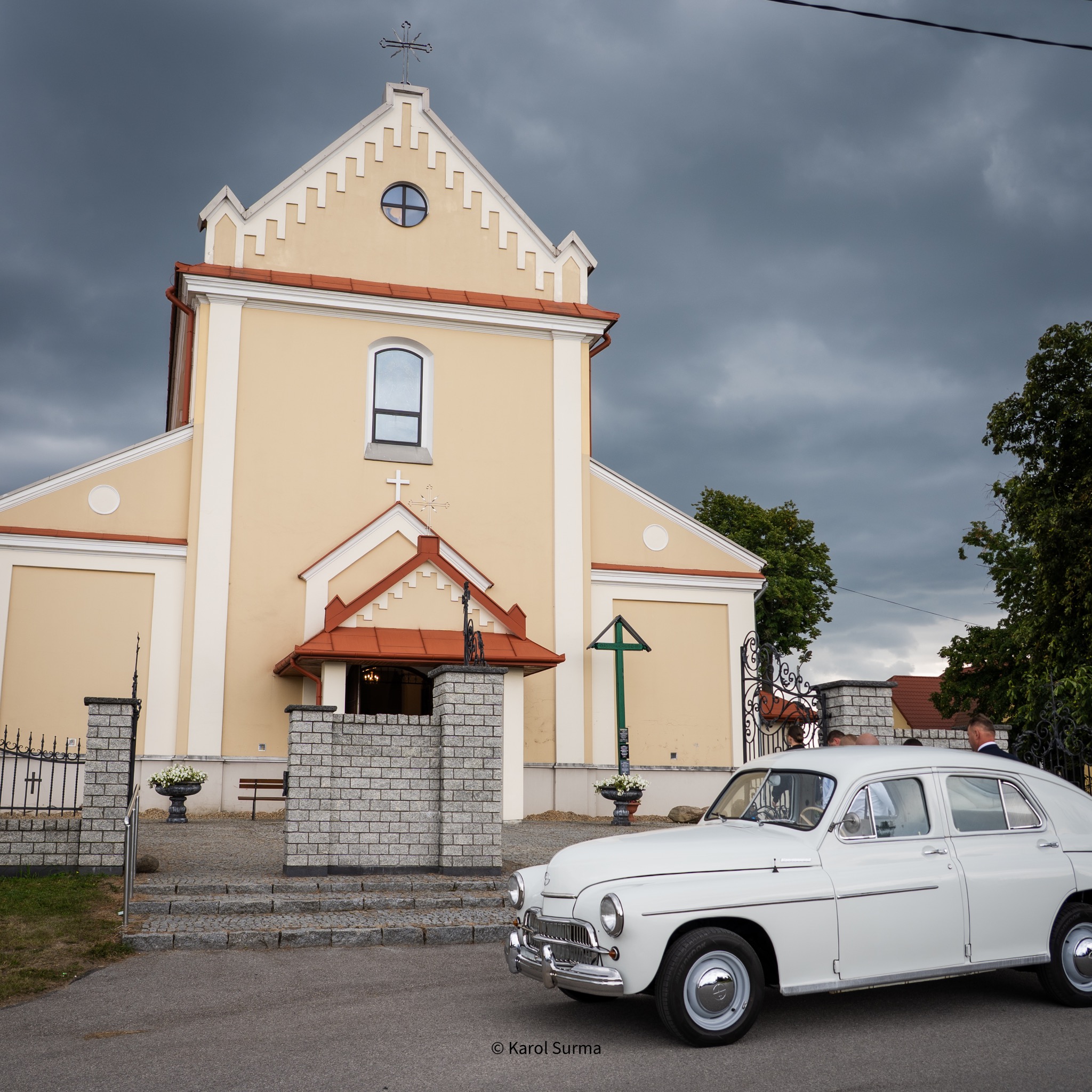 Biały zabytkowy samochód przed kościołem w Wadowicach, pochmurne niebo. Ujęcie z ceremonii ślubnej, detale architektury i pojazdu.