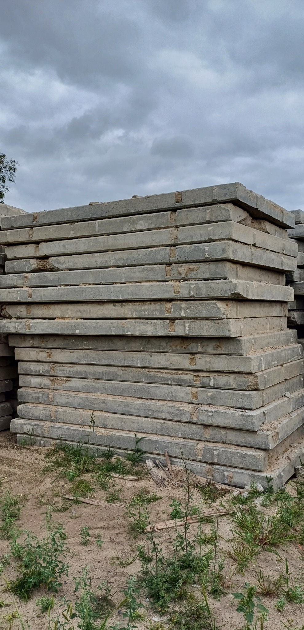 Stos szarych, betonowych płyt drogowych na piaszczystym gruncie, z widocznymi chwastami i pochmurnym niebem w tle. Płyty z widocznymi śladami użytkowania.