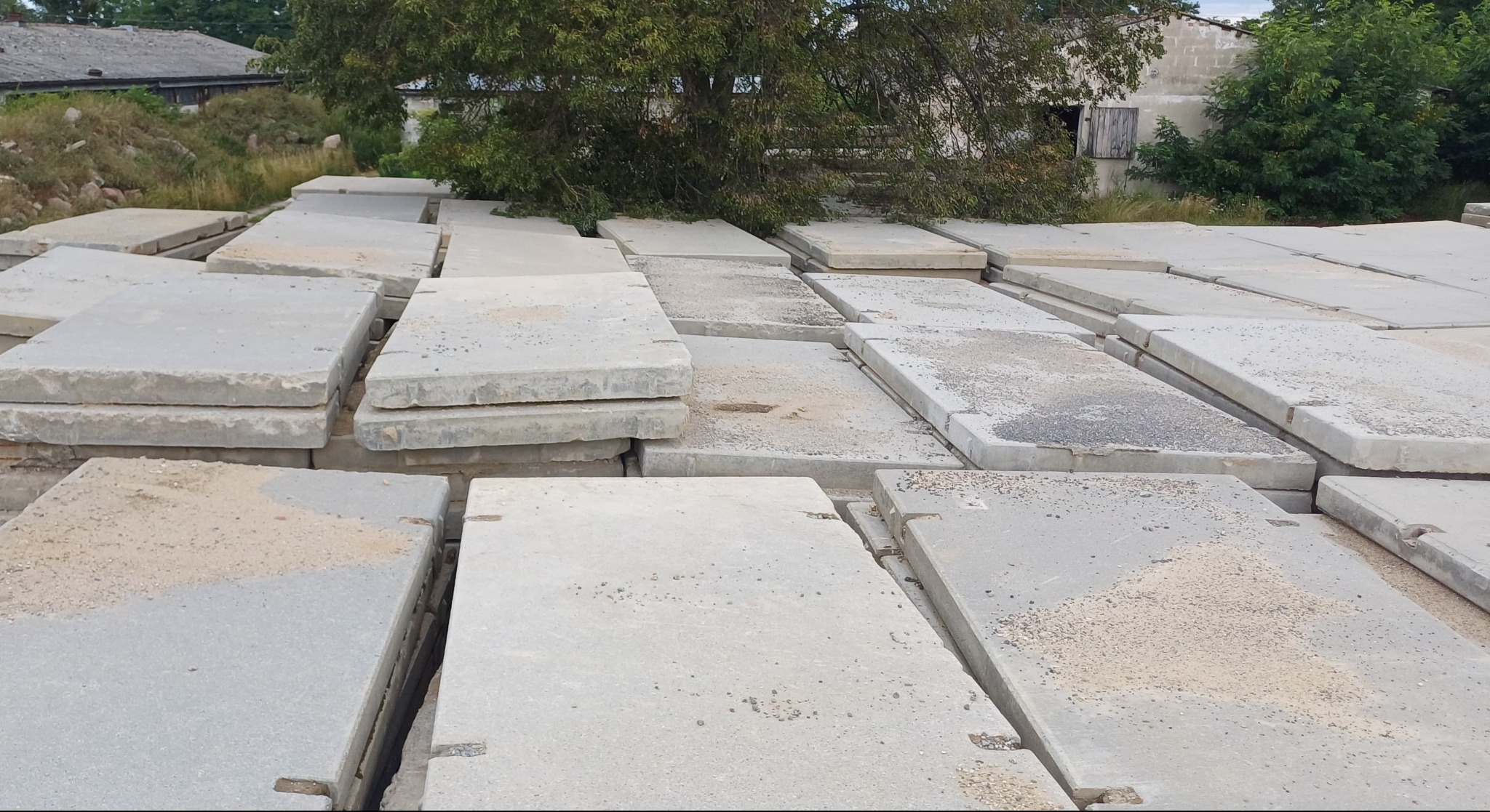 Szare płyty betonowe ułożone w stos, widoczne ślady piasku i ziemi, w tle budynki i zieleń. Płyty drogowe składowane na zewnątrz.