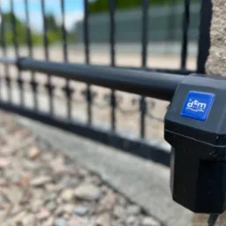 Czarny napęd automatyki bramy DTM, zamontowany na kamiennym słupku, z widoczną metalową bramą w tle i kamienistym podłożem.