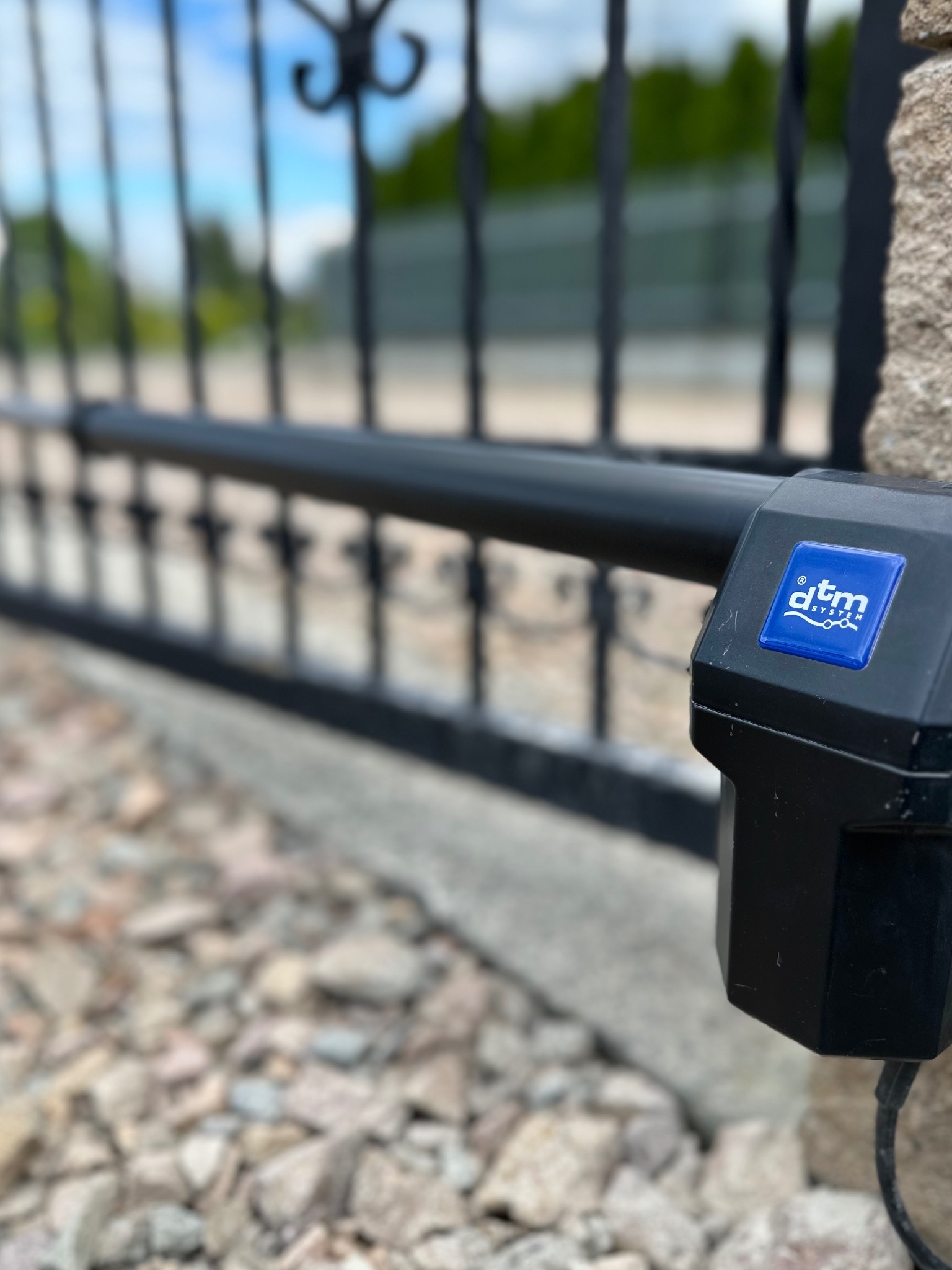Czarny napęd automatyki bramy DTM, zamontowany na kamiennym słupku, z widoczną metalową bramą w tle i kamienistym podłożem.