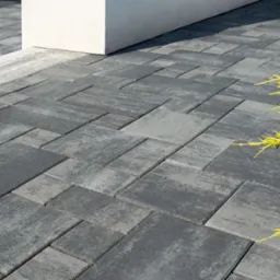 Ułożona kostka brukowa w odcieniach szarości, tworząca wzór. Widoczny fragment donicy i żółte źdźbła trawy. Ujęcie z bliska, podkreślające fakturę kostki.