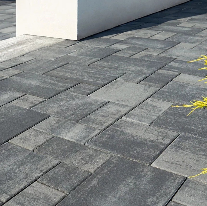 Ułożona kostka brukowa w odcieniach szarości, tworząca wzór. Widoczny fragment donicy i żółte źdźbła trawy. Ujęcie z bliska, podkreślające fakturę kostki.