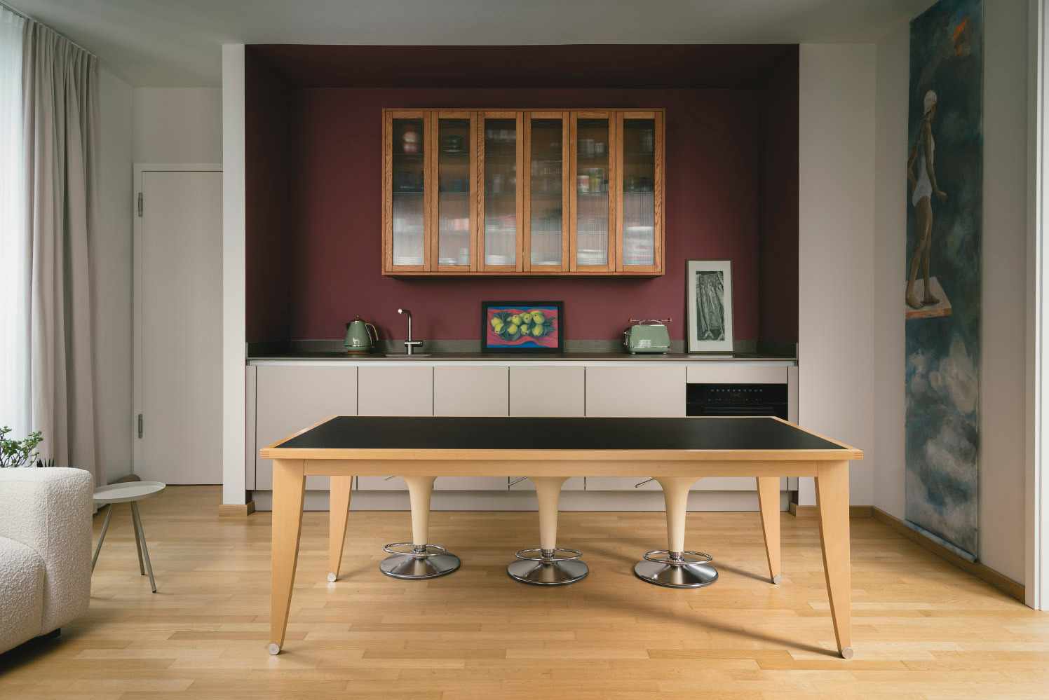 Nowoczesna kuchnia z drewnianymi szafkami, blatem w kolorze beżu i stołem z czarnym blatem. Na ścianie obraz z motywem pływaczki. Całość w ciepłej tonacji.