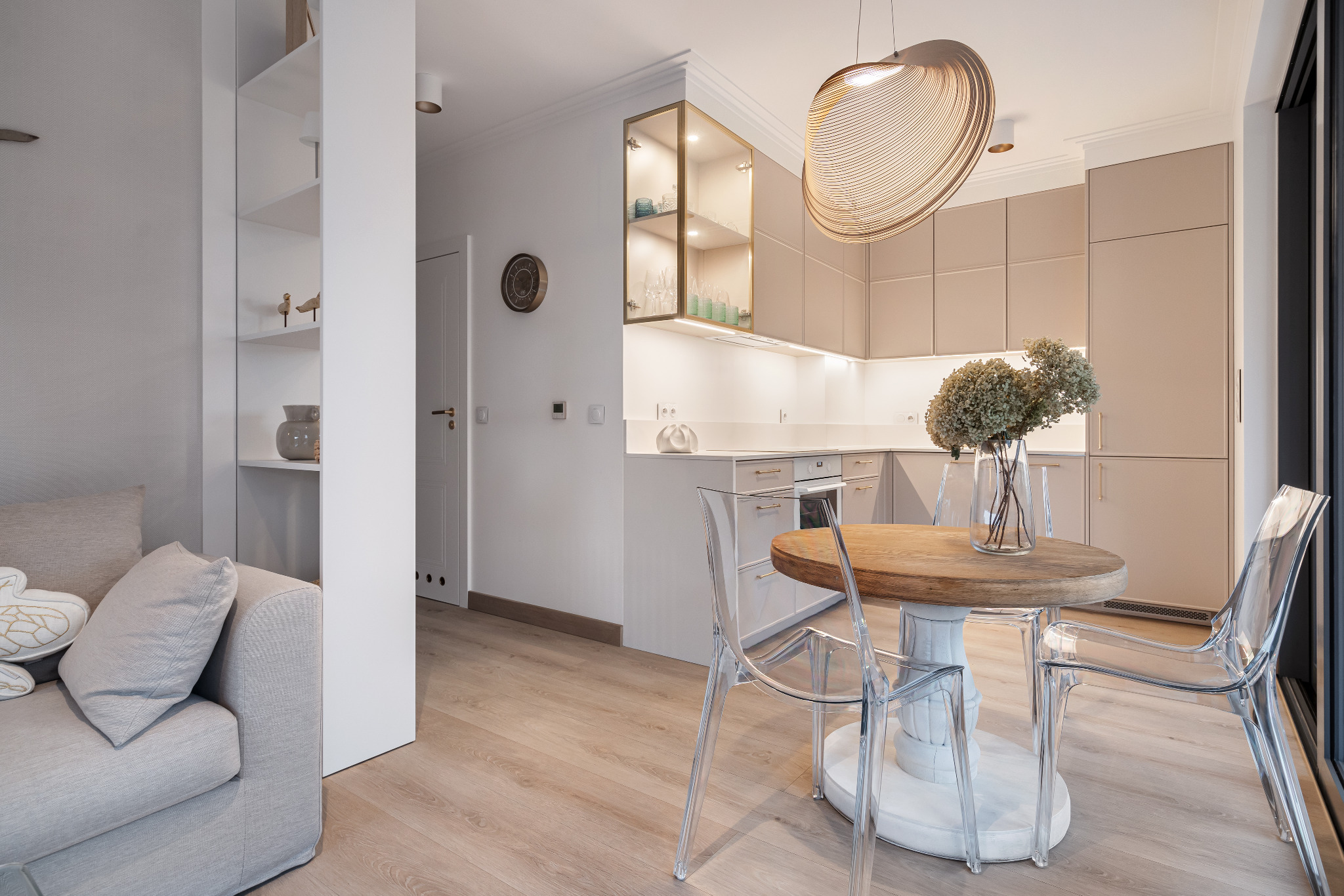 Jasne wnętrze: salon z kuchnią. Okrągły drewniany stół z przezroczystymi krzesłami, beżowe szafki kuchenne i białe półki. Nowoczesny wystrój.