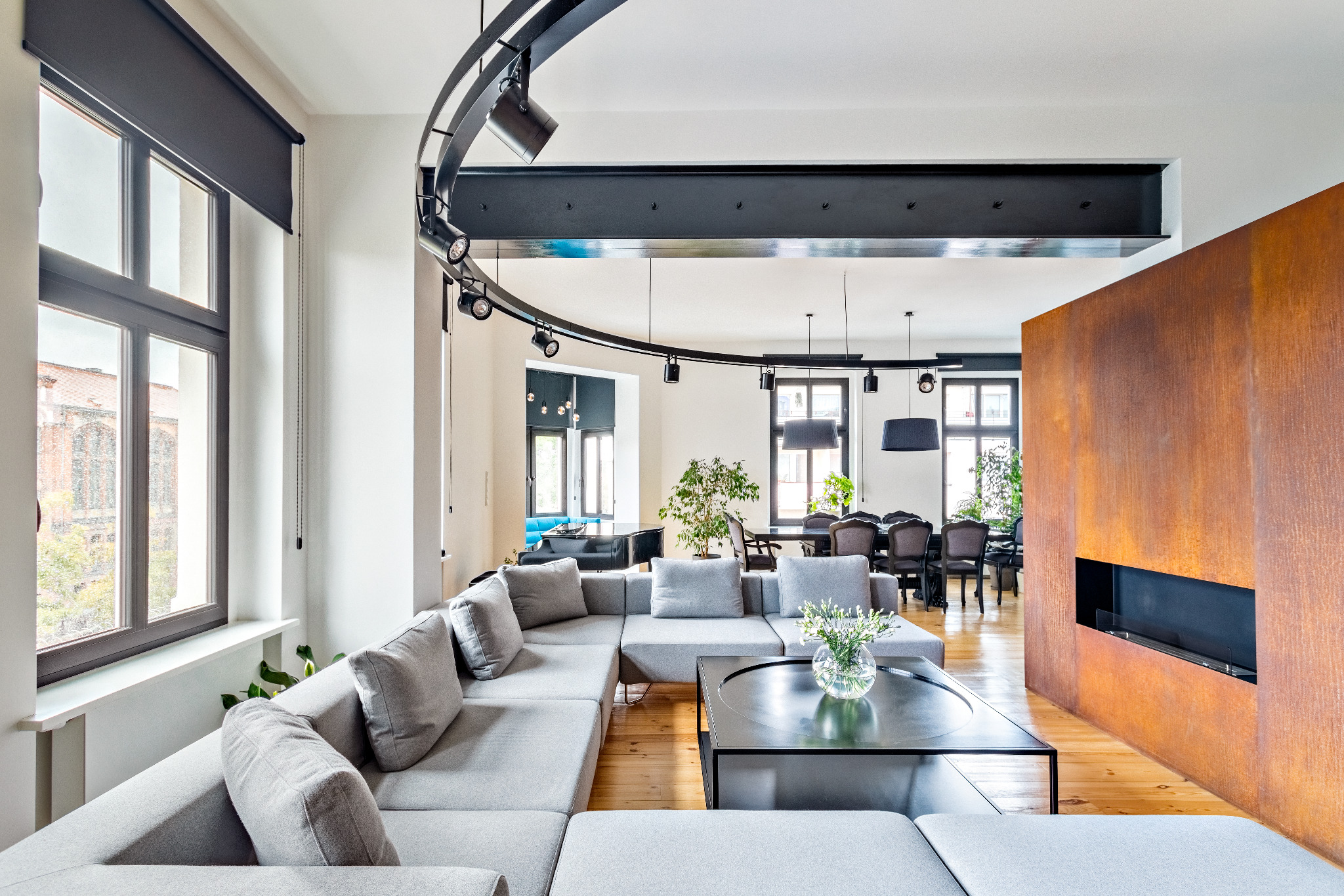 Nowoczesny salon z szarą sofą narożną, drewnianą podłogą i ścianą z efektem rdzy. Charakterystyczne oświetlenie sufitowe i widok na jadalnię w głębi.
