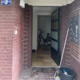 Otwarty korytarz wejściowy z rowerem w tle, wiadro z mopem przy ceglanej ścianie obok skrzynki pocztowej, numer domu 3.