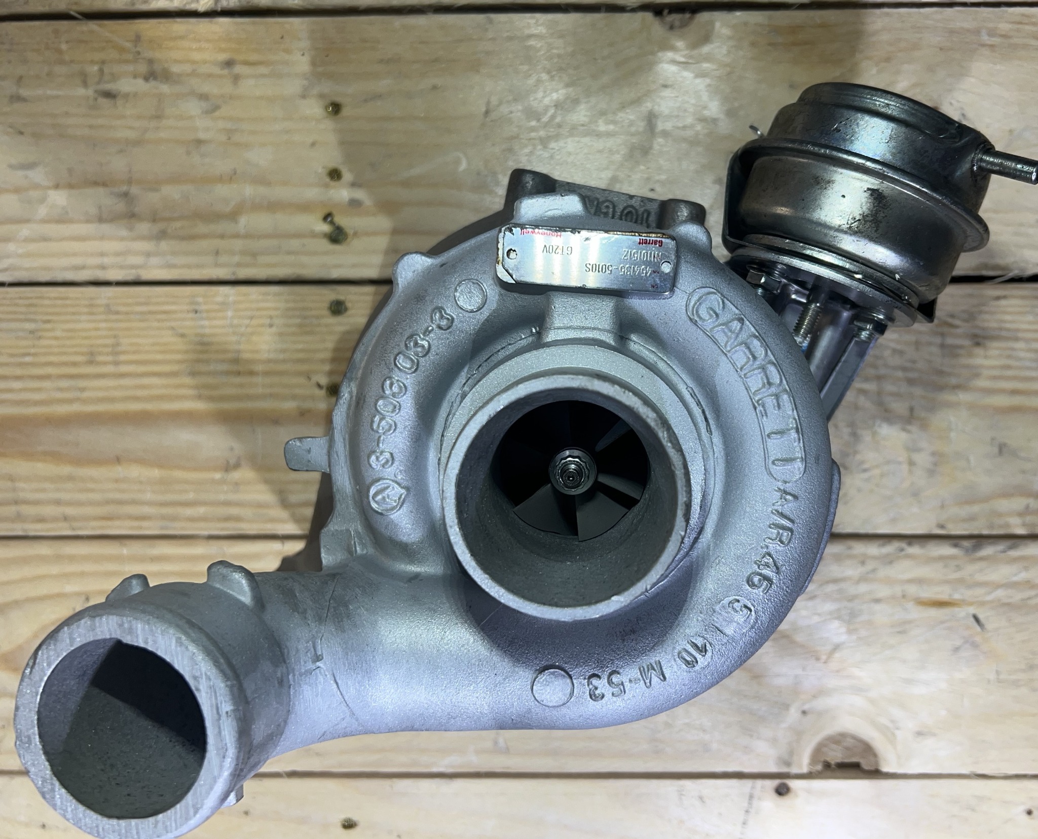 Szara turbosprężarka Garrett z widocznymi oznaczeniami modelu, leżąca na drewnianym podłożu. Widok z przodu na wirnik.