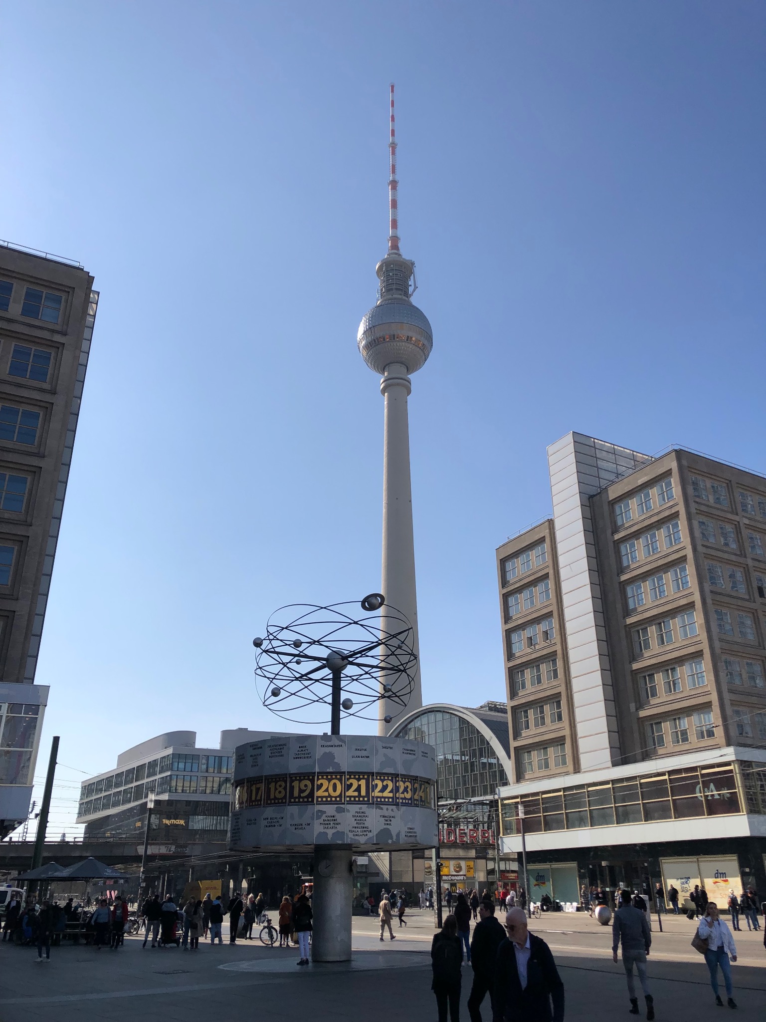 Panorama Alexanderplatz w Berlinie z wieżą telewizyjną i zegarem świata. Budynki w tle. Błękitne niebo. Dużo ludzi na placu.