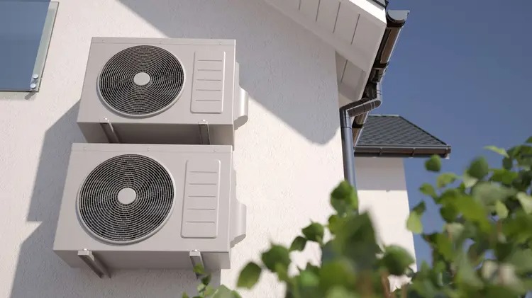 Dwa klimatyzatory na białej ścianie budynku, widoczne z dołu, z fragmentem dachu i rynny w tle oraz zielenią na pierwszym planie. Jasne, naturalne oświetlenie.