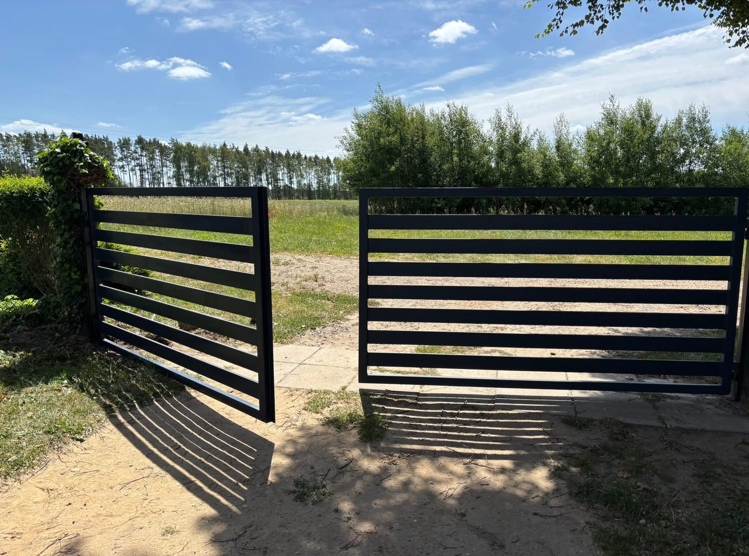 Nowoczesna, czarna brama wjazdowa z poziomymi szczeblami, otwarta na tle zieleni i błękitnego nieba. Cień bramy na piaszczystym podłożu.