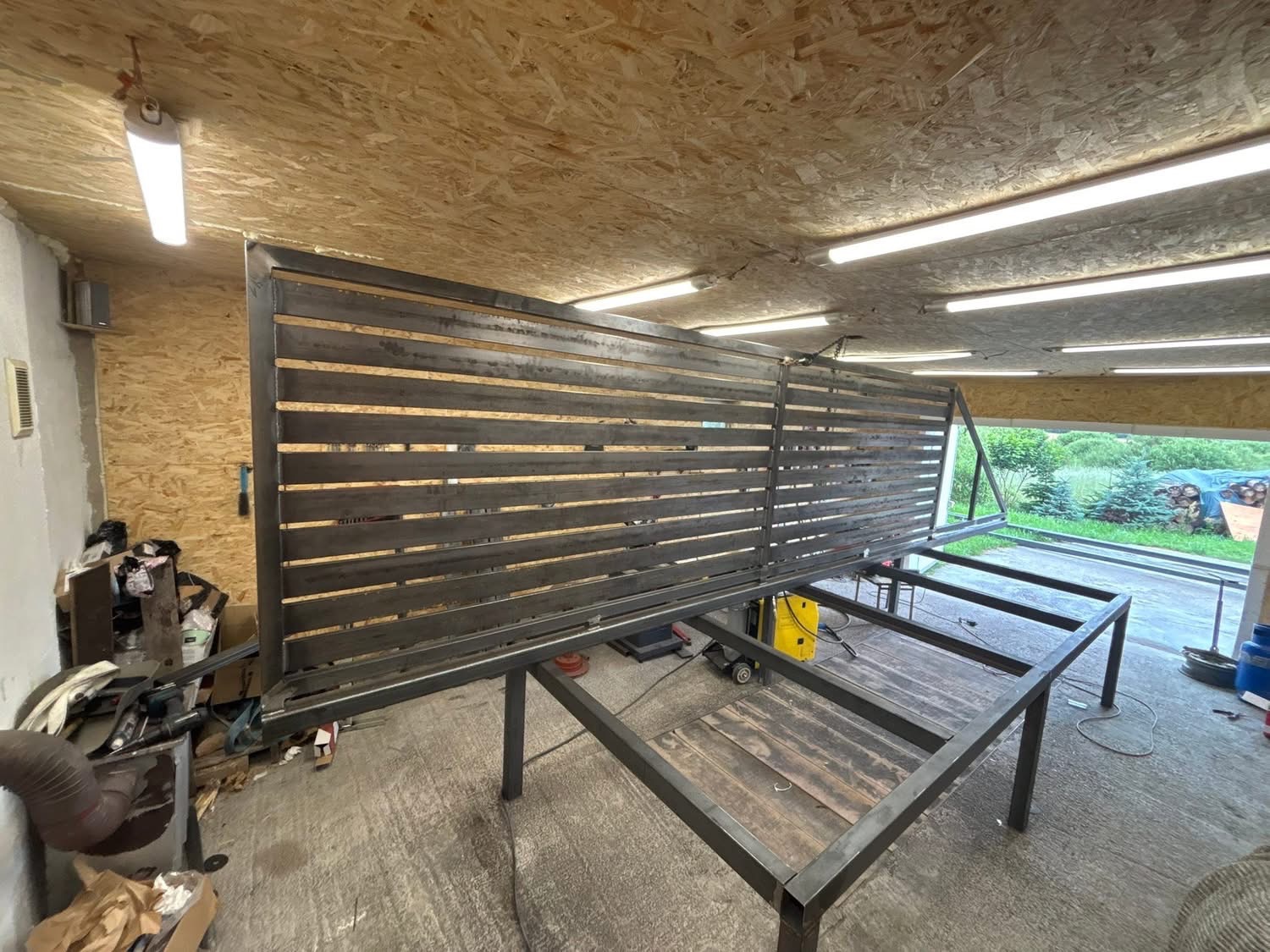 Konstrukcja bramy wjazdowej z poziomymi profilami, spawana w warsztacie. Widoczny stelaż i elementy mocujące, przygotowane do montażu. Surowy metal, oświetlenie warsztatowe.